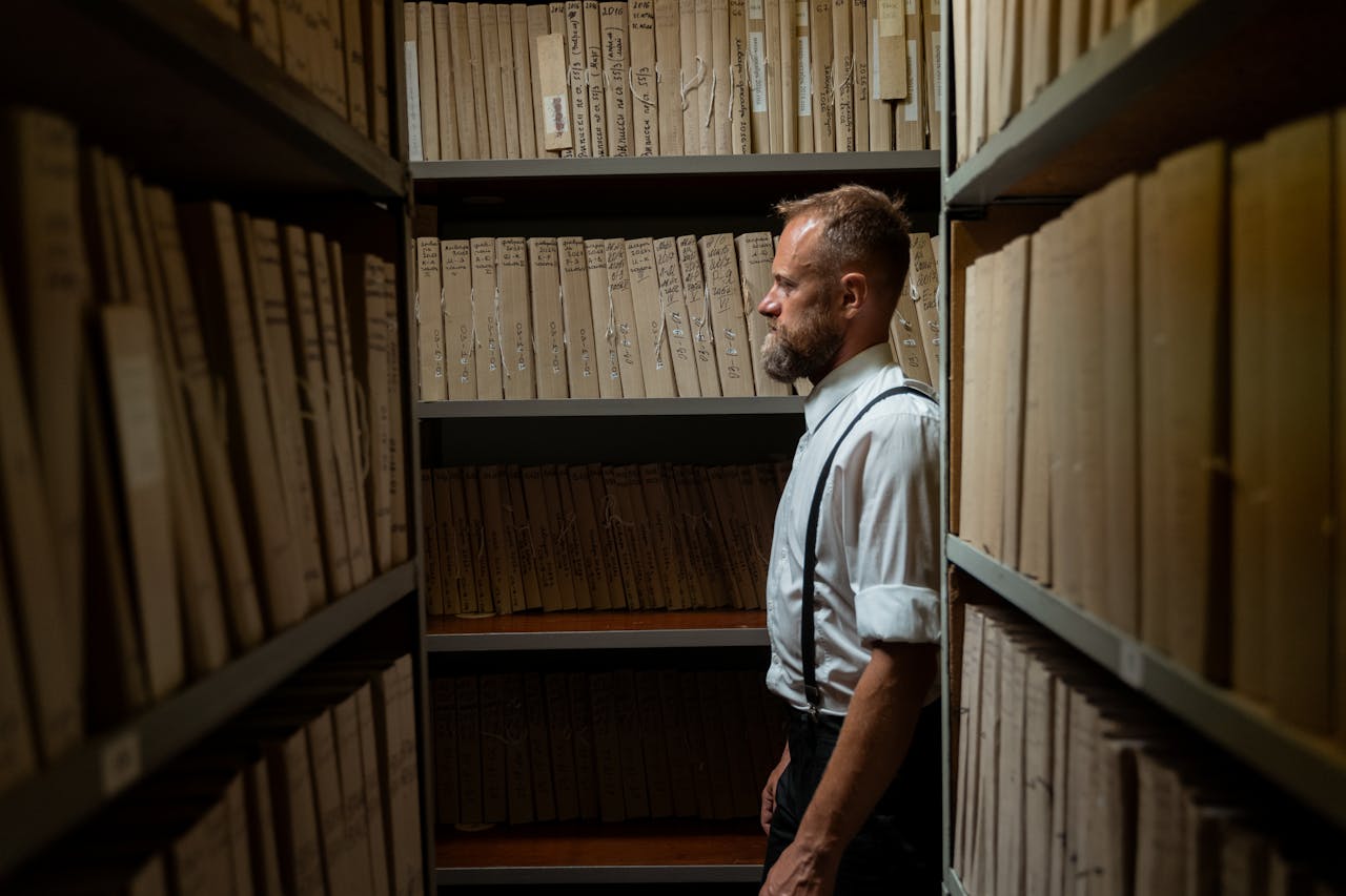 A man in formal attire and suspenders stands in an archive surrounded by documents.